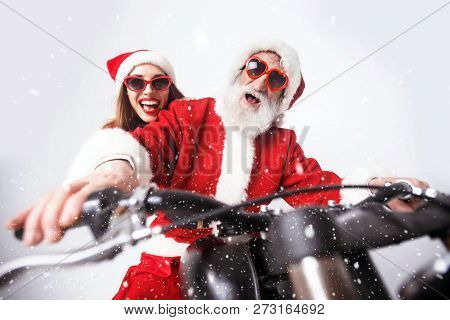 Santa Claus With White Beard Wearing Sungasses And Young Mrs. Claus Wearing Santa Hat, Red Sweater A
