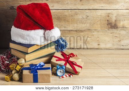 A Stack Of Books, Santa Clauss Cap, An Alarm Clock, Gift Boxes On A Wooden Background. Concept Of Ne