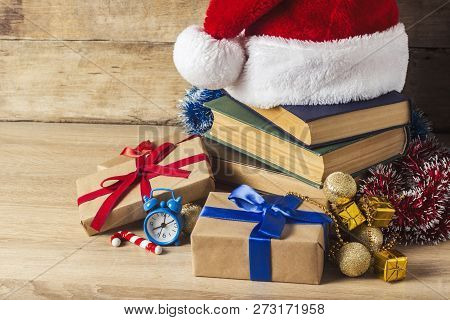 A Stack Of Books, Santa Clauss Cap, An Alarm Clock, Gift Boxes On A Wooden Background. Concept Of Ne