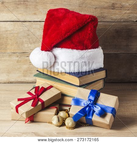A Stack Of Books, Santa Clauss Cap, Gift Boxes With Red And Blue Ribbons On A Wooden Background. Con