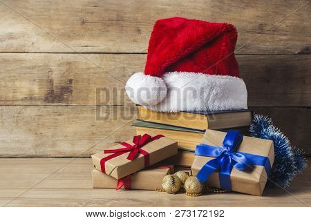 A Stack Of Books, Santa Clauss Cap, Gift Boxes With Red And Blue Ribbons On A Wooden Background. Con
