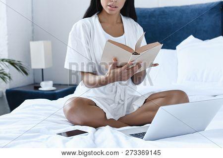 Cropped Image Of Mixed Race Girl In White Robe Reading Book On Bed In Morning