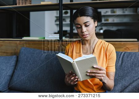 Beautiful Mixed Race Girl In Orange Shirt Reading Book On Sofa In Living Room