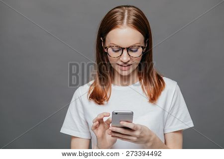 Portrait Of Pleasant Looking Relaxed Woman Holds Modern Cell Phone, Types Text Message On Cell Phone