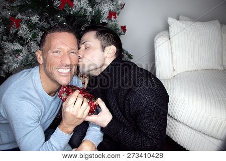 Gay Male Couple Exchanging Christmas Gift In Front Of Tree With A Kiss