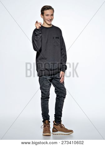 Teen boy is pointing by finger. Teenage boy dressed in black trendy clothes posing at studio. Full portrait of handsome teen boy over whitebackground.
