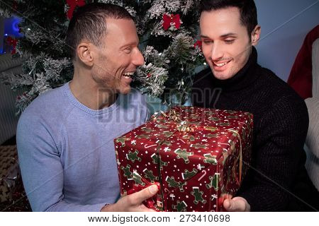 Happy Male Gay Couple Exchanging Christmas Gift In Front Of Tree With Red Ribbons