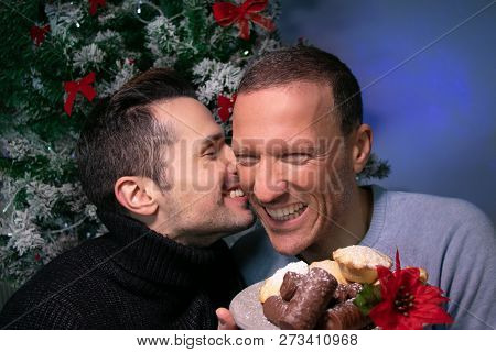 Male Gay Couple Kissing And Laughing In Front Of Christmas Tree With Plate Of Cakes