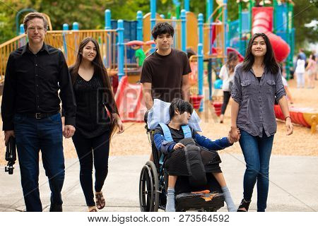Caucasian Father And Four Biracial Teen Children Walking Past Playground With Disabled Boy In Wheelc