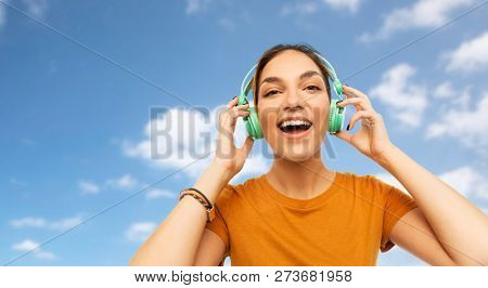 technology and people concept - happy young woman or teenage girl with headphones listening to music over blue sky and clouds background