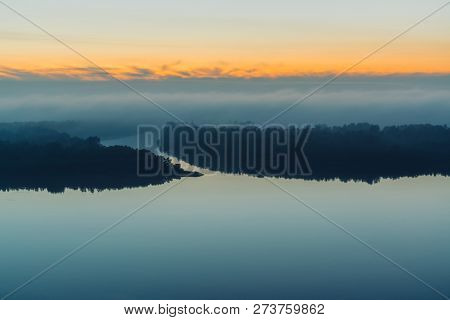 Broad River Flows Along Shore With Forest Under Thick Fog. Early Blue Sky Reflected In Water. Yellow
