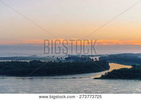 Early Blue Sky Reflected In River Water. Riverbank With Forest Under Predawn Sky. Orange Stripe In P