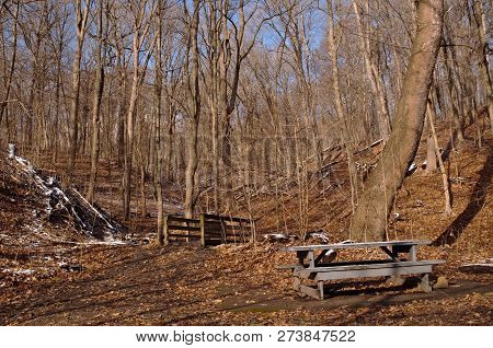 A Wooden Picnic Table Partially Covered In Snow Next To A Wooden Bridge In The Winter Woods In Frick