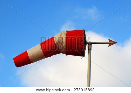 Windsock With A Weather Vane In Front Of A Blue Sky With White Clouds. The Windsock Is Red And White