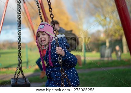 Pre-school Aged Caucasian Female In The Playground On The Swing, Smiling To Neutral Look, In The Bac