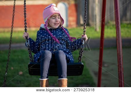 Pre-school Aged Caucasian Female In The Playground On The Swing Happy S,iling, Eyes Closed.