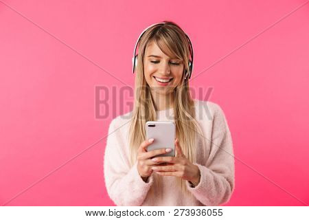 Cheerful young blonde woman wearing sweater standing isolated over pink background, listening to music with headphones, using mobile phone
