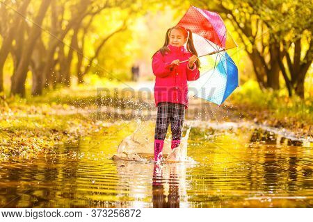 Little Girl In Rubber Boots Running Through A Puddle On A Rainy Day