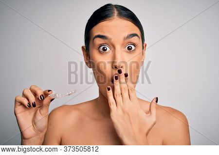 Young beautiful brunette woman holding dental aligner orthodontic to teeth corretion cover mouth with hand shocked with shame for mistake, expression of fear, scared in silence, secret concept
