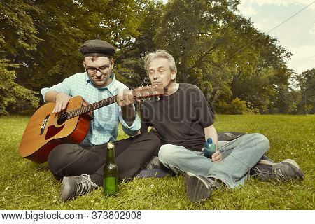 Two Men On City Park Summer Meadow Enjoying Day With Guitar