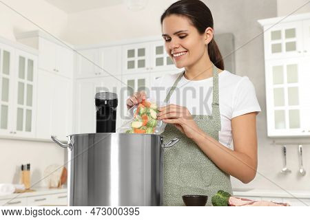 Woman Putting Vacuum Packed Vegetables Into Pot With Sous Vide Cooker In Kitchen. Thermal Immersion 