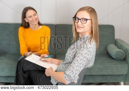 Smiling Female Psychologist With Glasses Looks Into The Camera, Consulting A Smiling Young Woman In