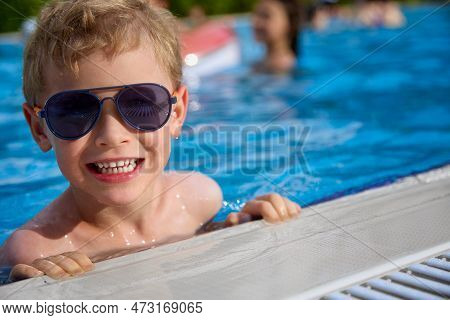Cute Child In Sunglasses Smiles, Holding On To The Side Of The Pool, Being In Warm Water. Fashionabl