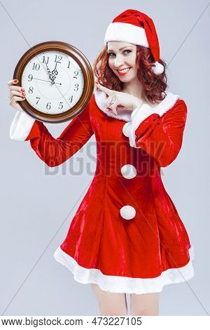 Winsome Gleeful Red-haired Santa Helper With Big Round Clock And Showing Time Posing Against White B