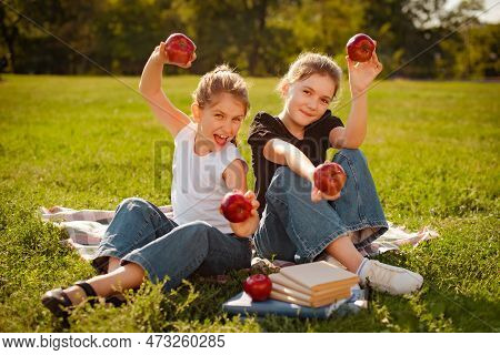 Two Girls Play With Red Apples On Green Grass In Park Or School Yard. Classmates Doing Homework Toge