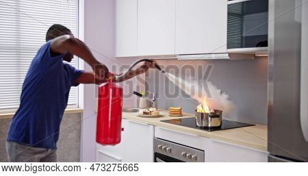 Man Using Fire Extinguisher To Stop Fire On Burning Cooking Pot In The Kitchen