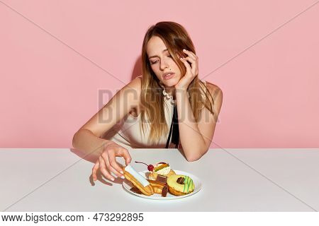 Adorable Girl Sitting At Table And Taking Ecler With Sad Facial Expression Over Pink Background