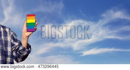 Female Hand Holding Smartphone With Lgbt Glag On The Background Of Cloudy Sky. Support, Stop Discrim