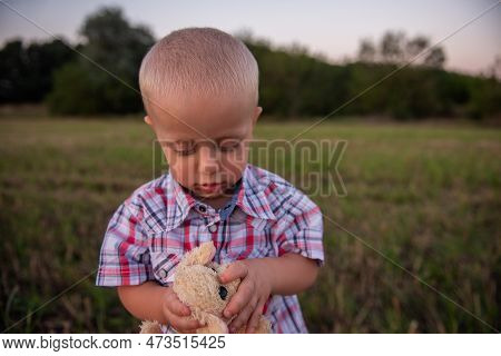 Little Boy In Cage Shirt Hugs Soft Toy. Todder With Emotions Walks In Mowing Green-yellow Field