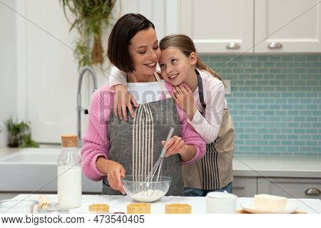 Glad Caucasian Young Mom And Little Daughter In Aprons Make Dough For Cookies, Hugs And Have Fun, En