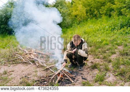 Front View Of Frozen Survivalist Male In Raincoat Putting Firewood On Campfire To Making Fire On Ban