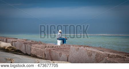 Port Aransas, Tx - 7 Feb 2023: Elderly Fisherman Sitting Patiently On A Plastic Pail While Fishing A