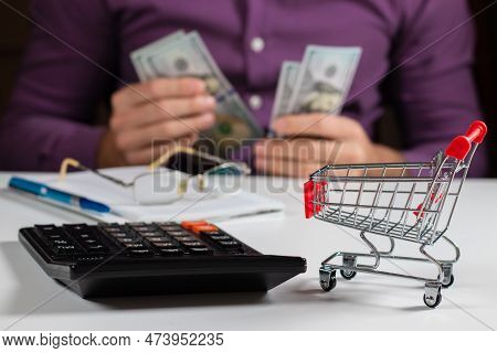Man Calculates The Budget. Shopping Cart A On Table With Calculator And Paper And Mohey In Hadns. Bu