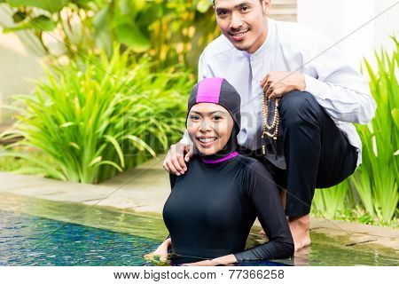 Muslim woman or girl in swimming pool in tropical garden wearing Burkini halal swimwear greeting her husband