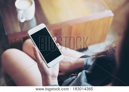Mockup image of a woman holding white mobile phone with blank black screen with coffee mug on wooden table in cafe