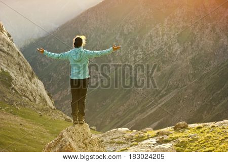 girl in blue jacket standing with hands up above mountain slopes at yellow sunset