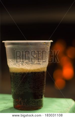 Dark beer (ale) in a plastic cup on the table with blurred bonfire lights background. Outdoor vacation concept.