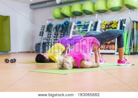 Group of women lying on floor on mats doing bridging exercise to firm buttocks indoors in fitness club.