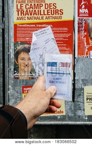 STRASBOURG FRANCE - APR 23 2017: French voter registration card held by male hand in front of official campaign poster of Nathalie Arthaud candidate for the 2017 French presidential elections posted outside a polling station