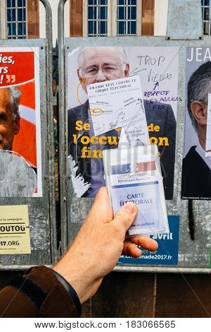 STRASBOURG FRANCE - APR 23 2017: French voter registration card held by male hand in front of official campaign poster of Jacques Cheminade candidate for the 2017 French presidential elections posted outside a polling station