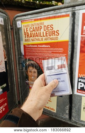 STRASBOURG FRANCE - APR 23 2017: French voter registration card held by male hand in front of official campaign poster of Nathalie Arthaud candidate for the 2017 French presidential elections posted outside a polling station