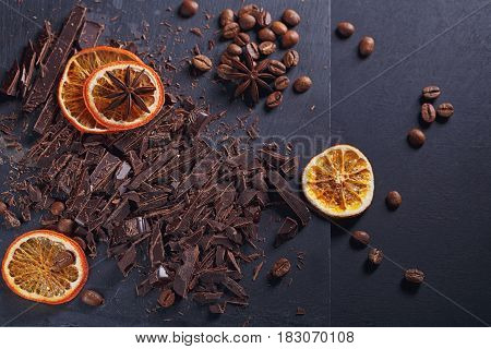 Dark chopping chocolate black roasted coffee beans dried lemon slices with spices anise on slate board over black background. Chocolate dessert confectionery concept. Top view with space for text.
