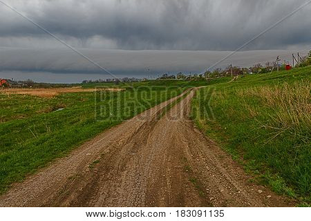 Dirt country road walking on a beam on the sides of which are new and last year's grass. In the distance are seen the houses of the village.In the sky hung a massive freakish forms of the clouds create a General dark background of the image.