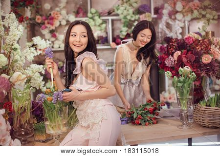 Beautiful asian florist girls making bouquet of flowers on table for sale against floral bokeh background in flower shop indoors. Two attractive asian females florists working in store. 2 playful fashion models in tender dresses happy smiling and playing 