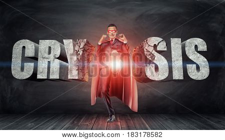 A businessman in a red cape and a mask kicking concrete letters reading 'Crysis' with his foot. Overcoming difficulties. Business and competition. Financial troubles.