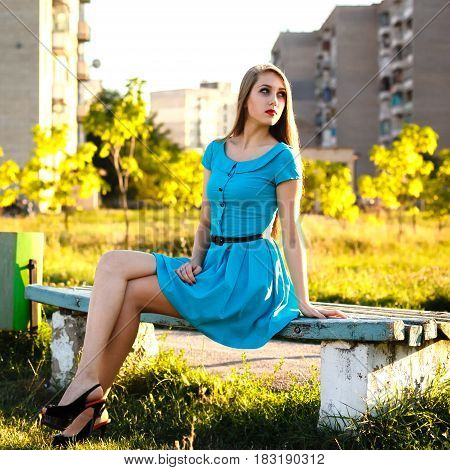 Beautiful young woman with long blonde hair sitting on a bench in a city park at sunny sammer day. Backlight.